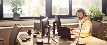 En mann som sitter og jobbed på data ved et skrivebord i et kontor med store vindu og noen planter i vinduskarmen